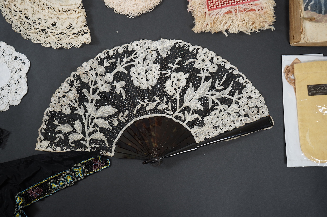 A French tortoiseshell, net, sequin and Honiton bobbin lace fan, with a collection of Jet appliques, a bead work fronter, embroidered mats and silk mats, an early Tiffany &Co poshette and embroidered appliqués and a Le M